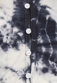 Niebiesko-kremowa tkanina w stylu tie-dye o miękkiej fakturze. Z przodu znajdują się pięć białych guzików, zdobionych kontrastowym przeszyciem wzdłuż plisowania.