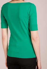 Kvinna iförd en grön skjorta med ärmar till armbågarna och svarta byxor, visad bakifrån mot en enkel ljus bakgrund.