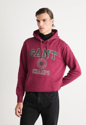 Burgunderfarget hettegenser med grønn brodert tekst "GANT CHAMPS" og et sirkulært logo. Har en lommesekk foran og snor i hetta. Brukt med svarte jeans.