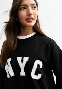 Sudadera negra con letras en blanco "NYC" en negrita, cuello redondo con ribete blanco, corte holgado y textura de tela suave.