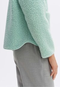 Pullover in pile verde menta chiaro con superficie testurizzata, orlo arrotondato e vestibilità rilassata, abbinato a pantaloni in tessuto grigi.