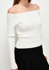 Weißes gerippte Off-Shoulder-Oberteil mit langen Ärmeln und taillierter Silhouette, kombiniert mit einem schwarzen plissierten Rock.