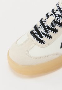 Sneaker med en kremfarget syntetisk overdel og beige semsket skinn-detaljer, med tykke svarte og hvite snører, og en gummisåle.