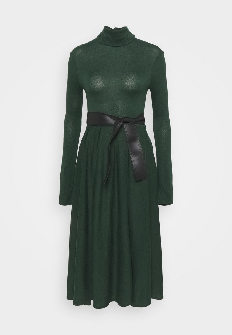 Robe midi vert foncé à manches longues avec col roulé et une large ceinture noire nouée à la taille sur un fond uni.