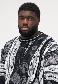 Black and white patterned sweater featuring intricate designs, varying textures, and a ribbed neckline. Heavy knit material with a relaxed fit.