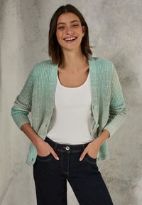Ljus mintgrön stickad cardigan med en texturerad yta, med knappstängning och ribbade ärmslut. Bärs över en vit topp och mörkblå jeans.