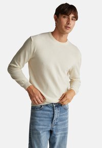 Lichtbeige sweater met lange mouwen en een ronde hals, gemaakt van zachte stof, gecombineerd met blauwe jeans. Eenvoudig ontwerp en getailleerde pasvorm.