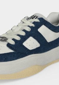 Basket décontractée blanche et bleue avec des lacets blancs, une semelle épaisse crème et un petit logo en métal près de la zone des orteils.