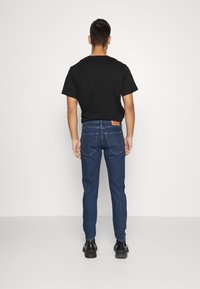 Camiseta negra de hombre combinada con jeans azules, con un diseño ajustado, cinco bolsillos y una etiqueta roja en la cintura trasera.