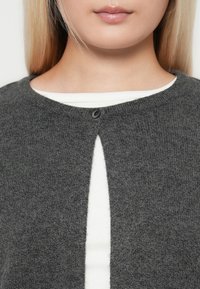 Vue partielle d'une personne portant un cardigan en tricot gris fermé par un seul bouton sur un haut à rayures blanc.