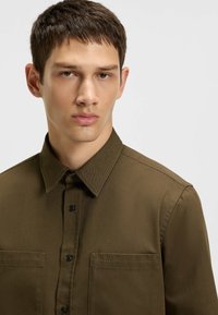 Brown button-up shirt with a collar, featuring a textured fabric, black buttons, and a small "HUGO" logo on the left chest pocket.