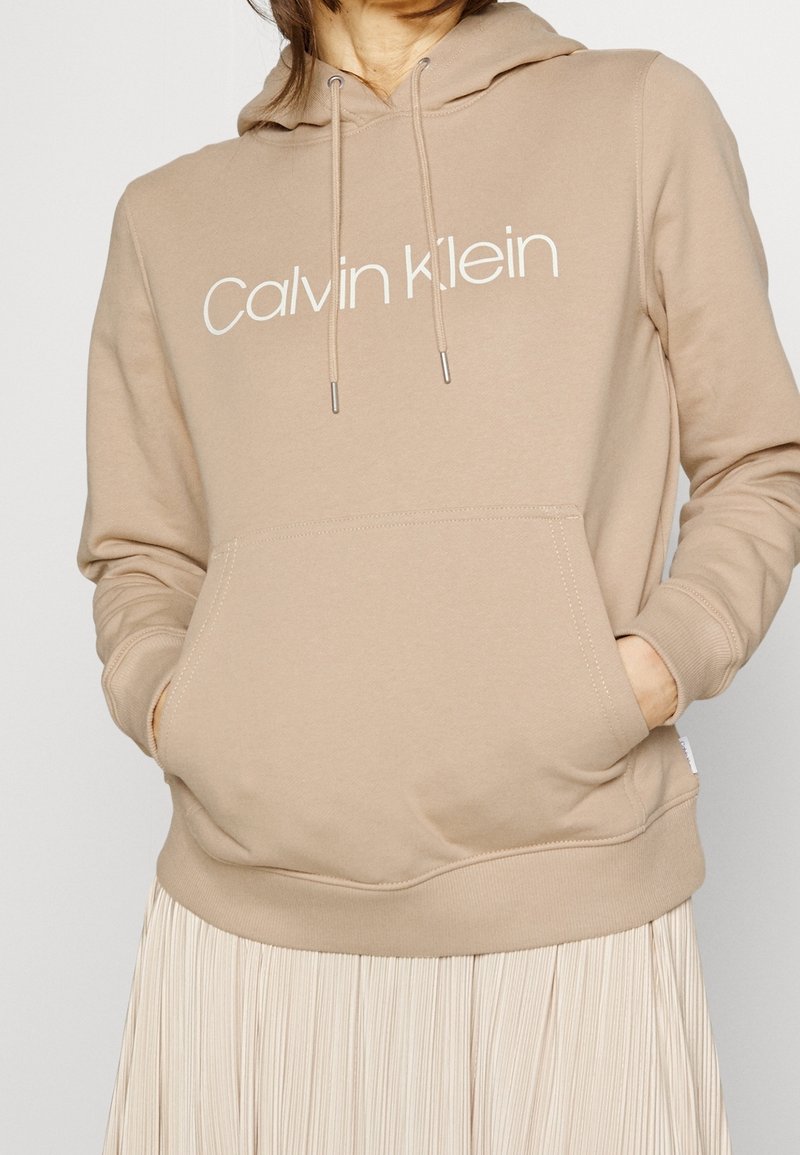 Béžová mikina s kapucí, přední kapsou a stahovací šňůrkou. Na mikině je bílé logo "Calvin Klein". Měkká tkanina, pohodlný střih, kombinována s plisovanou sukní.