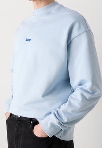 Felpa azzurra a girocollo con colletto e polsini a costine, caratterizzata da un patch rettangolare blu sul davanti, indossata sopra pantaloni neri.
