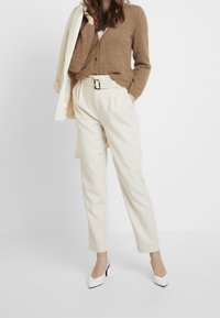 Ljusbrun stickad cardigan, vit skjorta, krämfärgade avsmalnade byxor med bälte och vita spetsiga klackskor mot en neutral bakgrund.