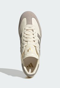 Zapatilla deportiva con un upper de cuero beige, acentos de ante gris, tres rayas grises y detalles de logo dorado en la lengüeta. Cordones redondos.