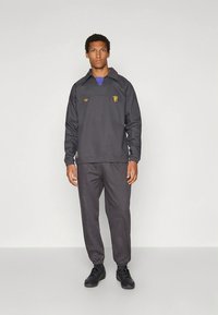 Σκοτεινό γκρι φόρμα που περιλαμβάνει μπλούζα με γιακά και τσέπη μπροστά, με ελαστικά μανσέτα, σετ με αντίστοιχο στενό παντελόνι και μαύρα αθλητικά παπούτσια.