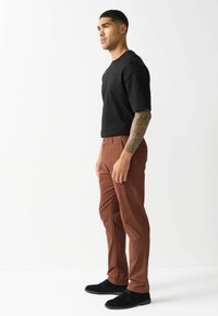 Schwarzes Kurzarmhemd und braune schmal geschnittene Hose, getragen mit schwarzen Schuhen. Das Hemd hat einen Rundhals; die Hose hat Seitentaschen und eine glatte Textur.