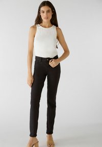 Zwarte slim-fit jeans met een klassiek vijf-pocketontwerp, gecombineerd met een witte ribbel tanktop. Model staat op beige blokhakken sandalen.