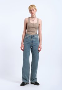 Junge Person mit kurzen blonden Haaren trägt ein beiges Tanktop, hoch taillierte blaue Jeans, schwarze Schuhe und geschichtete Halsketten und steht vor weißem Hintergrund.