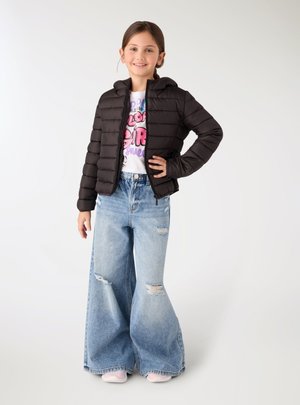 Ragazza sorridente in piedi con la mano sul fianco, indossa una giacca imbottita nera, una t-shirt bianca con una grafica, jeans larghi strappati e scarpe rosa su sfondo bianco.