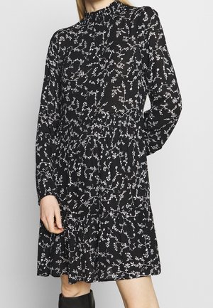 Frau trägt ein schwarzes, langärmeliges Kleid mit weißem Blumenmuster und hohem Kragen, posiert mit einer Hand an der Seite.