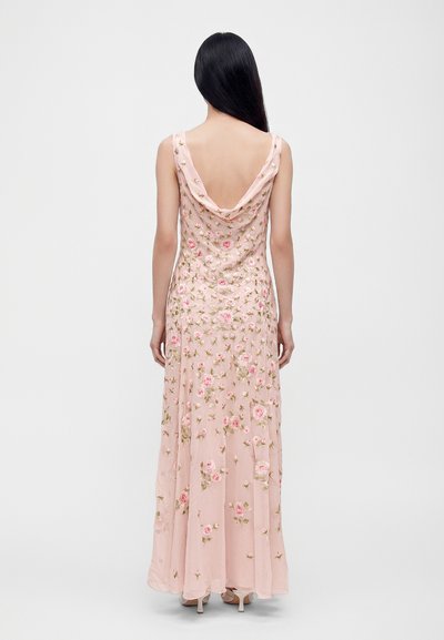 Femme aux longs cheveux foncés portant une robe longue rose à motifs fleuris avec un dos drapé et décolleté, et des talons hauts beiges, debout devant un fond blanc.