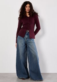 Burgundy top s dlouhým rukávem a zapínáním na knoflíky, s peplum lemem, kombinovaný s modrými džínami širokého střihu, které mají vybledlou texturu a poutka na opasek.
