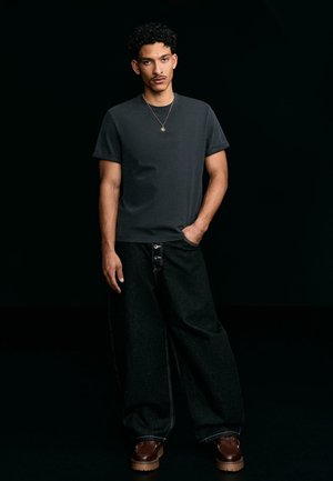 Jeune homme aux cheveux bouclés portant un t-shirt gris foncé, un pantalon large noir, des chaussures marron et un collier, debout devant un fond noir.