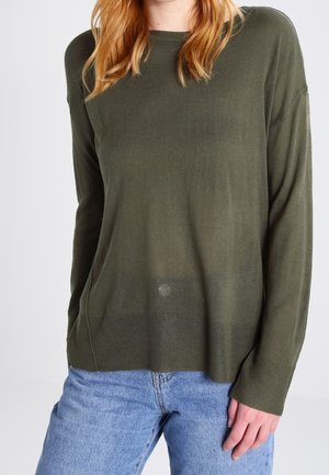 Femme portant un pull à manches longues vert olive et un jean bleu clair, debout avec une main près de sa hanche sur un fond uni.