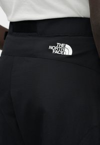 Κοντινό πλάνο προσώπου που φοράει μαύρο παντελόνι North Face με λογότυπο στο πάνω μέρος του γοφού, σε συνδυασμό με λευκό τοπ.