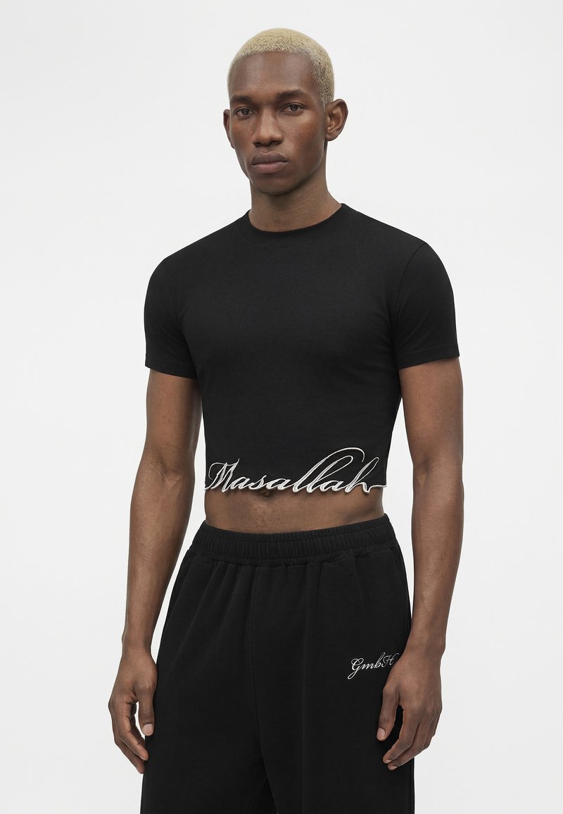 Young man with short blonde hair wearing a black cropped t-shirt with "Masallah" text and black pants with white logo on left thigh.