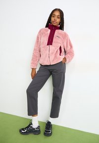 Fluffy roze fleecejas met bordeauxrode accenten, rits aan de voorkant en zijzakken. Gecombineerd met grijze broek en zwarte sneakers. Groene vloer achtergrond.