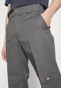Personne portant un pantalon de travail gris avec des poches latérales, une main dans la poche, et un patch logo de la marque Dickies visible sur la partie inférieure de la jambe.