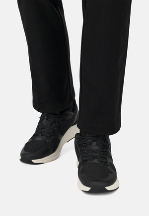 Zapatos deportivos negros con parte superior de malla y sintético, con cordones, cuello acolchado y una entresuela de color claro. Llevados con pantalones negros.