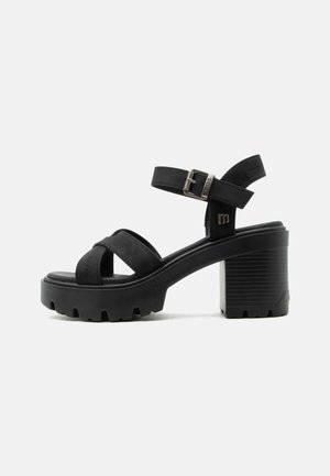 Sort chunky hælsandal med krydsede remme, ankelspænde og rillesål på en ensfarvet hvid baggrund.