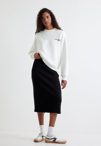 Witte sweatshirt met tekst, gecombineerd met een zwarte midi rok. Model draagt witte sneakers met zwarte accenten en witte sokken tot aan de enkel.