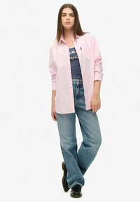 Jasnoróżowa koszula na guziki z podwiniętymi rękawami, noszona na ciemnoniebieskim T-shircie z nadrukiem. Dopasowana do luźnych dżinsów i czarnych butów.