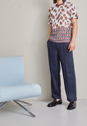 Hvid skjorte med korte ærmer med røde blomster- og geometriske mønstre; marineblå bukser med vide ben; sorte slip-on sko; lyseblå lænestol i nærheden.