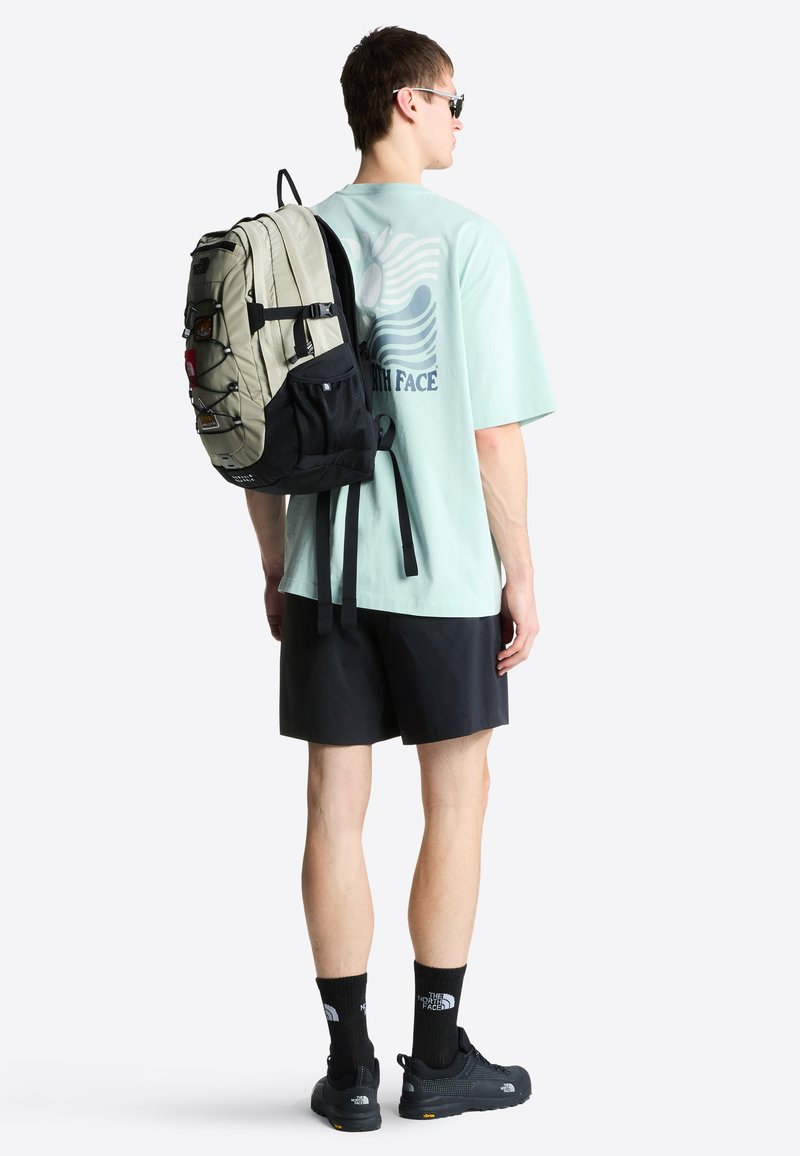 Mann trägt Sonnenbrille, mintgrünes T-Shirt mit Grafik, schwarze Shorts, Socken und Schuhe, und hat einen beige-schwarzen Rucksack auf einer Schulter getragen.