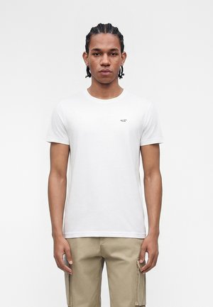 Junger Mann mit geflochtenem Haar, der ein schlichtes weißes T-Shirt mit kurzen Ärmeln und beige Cargohosen trägt, steht vor einem weißen Hintergrund.