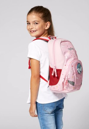 Lächelndes Mädchen, das ein weißes T-Shirt und blaue Jeans trägt, und einen rosa Rucksack mit roten Riemen und einem verspielten Einhorn- sowie Punkte-Design trägt.