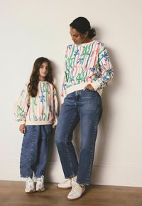 Zwei passende Sweatshirts in Pastellfarben mit grünen und blauen Blumenmustern. Die Mutter und das Kind tragen Jeans und helles Sneaker.