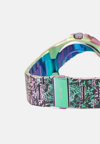 Bracelet de montre en métal avec des maillons multicolores et irisés aux motifs ornés. Boucle verte gravée "GUESS" avec des détails complexes.