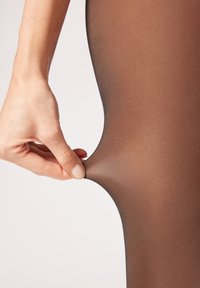 Braune, durchsichtige Strumpfhose, die mit den Fingern gezogen wird und eine glatte Textur sowie ein leichtes Material zeigt. Einfaches, sauberes Design, frei von Mustern.