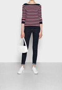 Marine gestreifter Pullover mit rot-weißen Streifen, dunkle Slim Jeans, weiße steppierte Handtasche und weiße Sneaker. Lässiges Outfit vor neutralem Hintergrund.