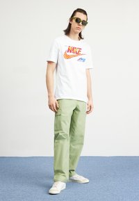 Hvid T-shirt med rød og gul "Nike" grafik; grøn cargo-bukser med sidelommer; hvide sneakers; solbriller.
