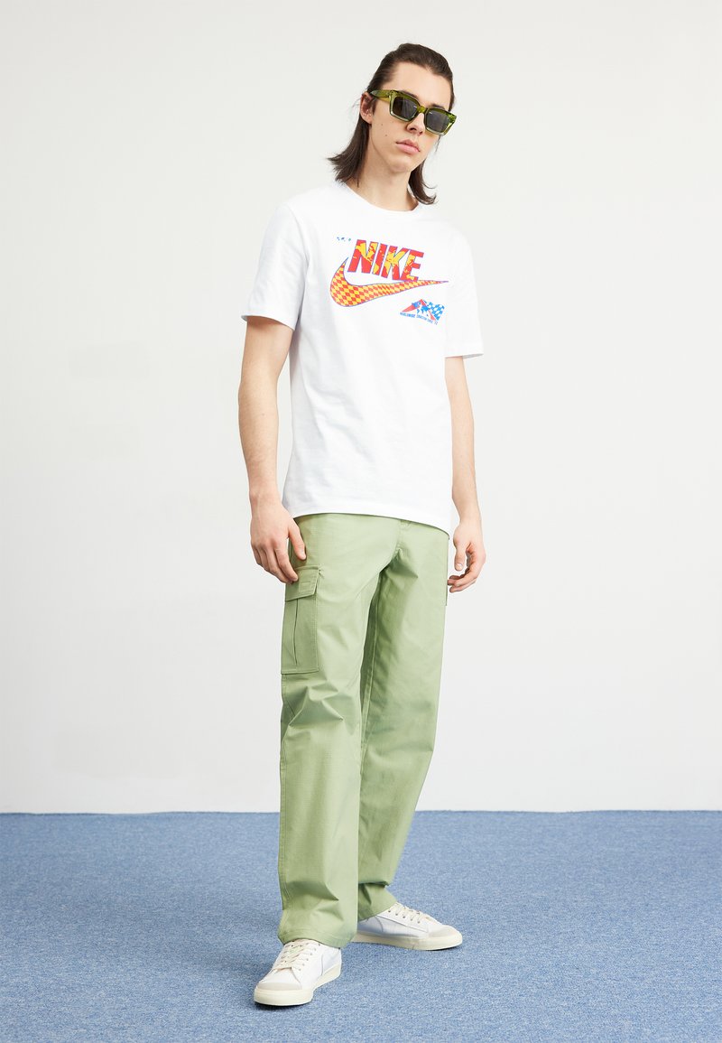 T-shirt blanc avec un graphisme "Nike" rouge et jaune ; pantalon cargo vert avec poches latérales ; baskets blanches ; lunettes de soleil.