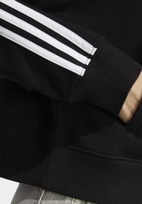 Svart ärm med vita trippelränder och ribbad mudd, hand i fickan på ett svart plagg, ljusa byxben synliga längst ner.