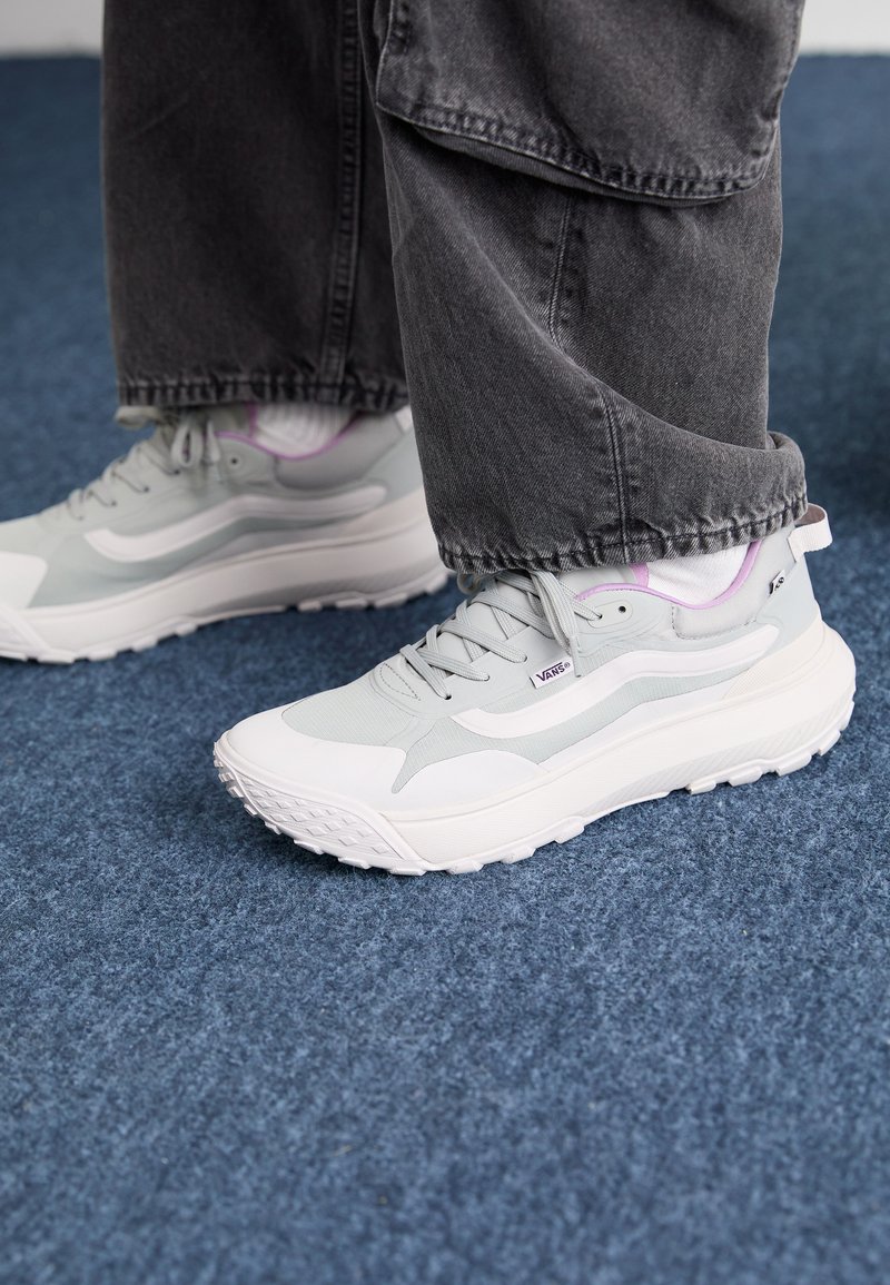 Persona con zapatillas Vans ligeras grises y blancas con acentos morados, usando jeans oscuros, de pie sobre una alfombra azul.