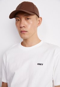 Brun hatt med buet skjerm, laget av glatt materiale. Hvit T-skjorte med en liten svart "OBEY"-logo på brystet.